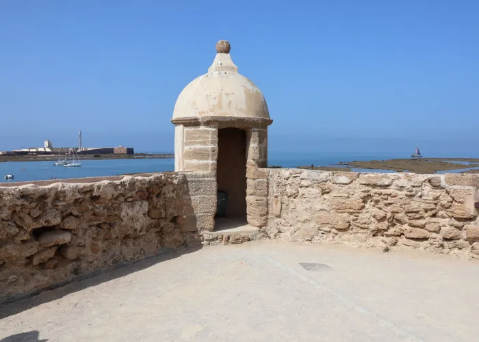 A historic stone watchtower overlooking the serene coastline of Cádiz, Spain, under a clear blue sky.
