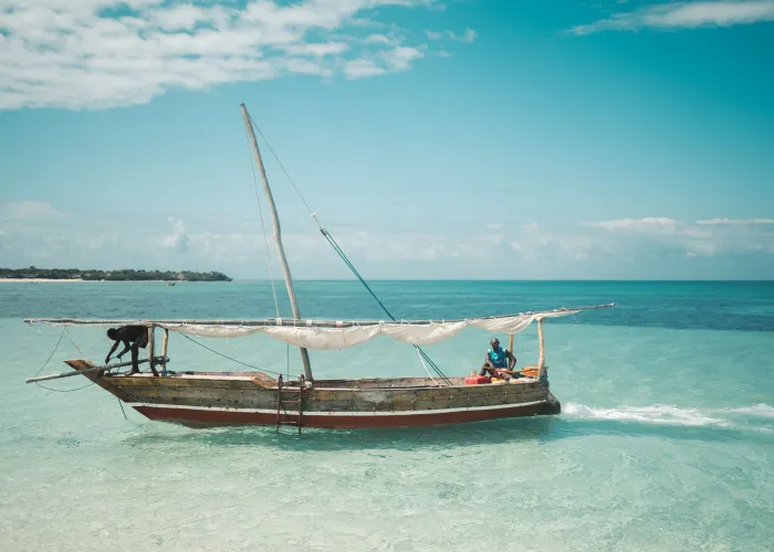 Two local fishermen navigate a traditional dhow on the clear waters of Zanzibar under a bright blue sky.