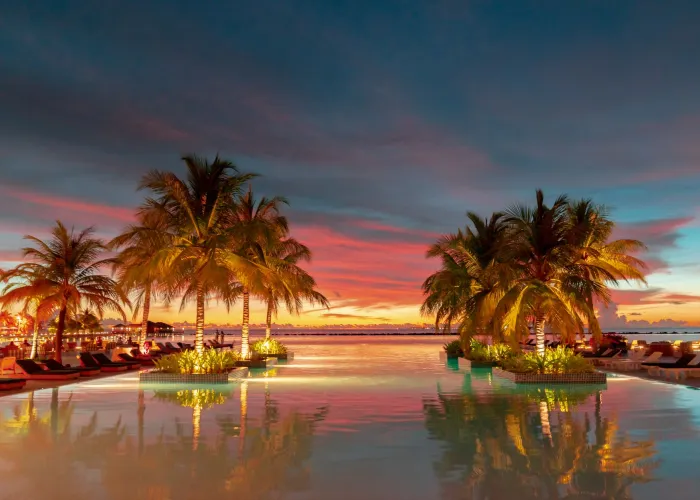 Beautiful sunset over a tropical resort pool with palm trees and vibrant sky reflections.