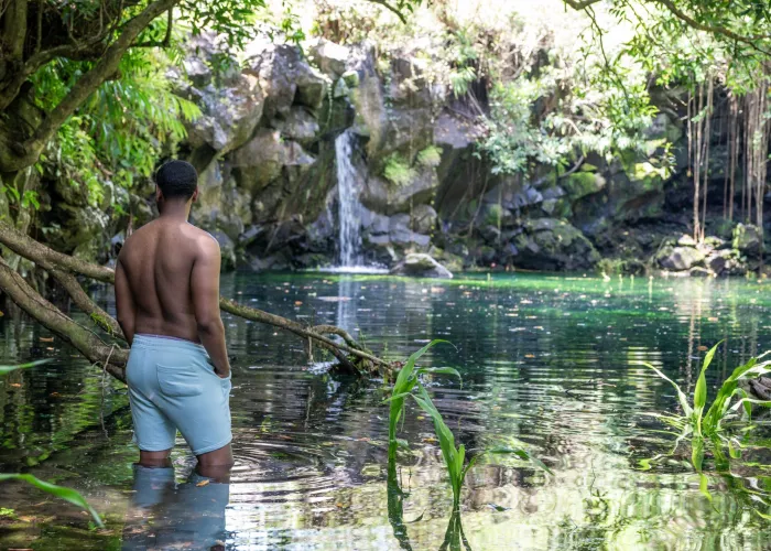A man stands in a tranquil pond surrounded by lush greenery with a waterfall in Mauritius.
