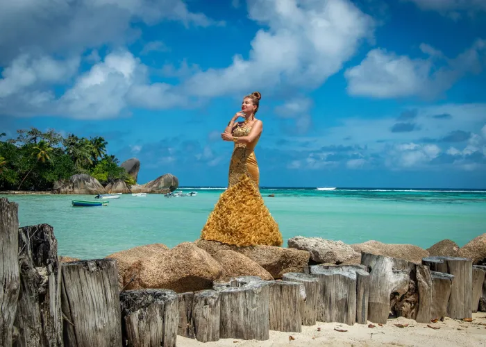 A woman in a golden dress poses elegantly by the turquoise sea on a sunny tropical beach.